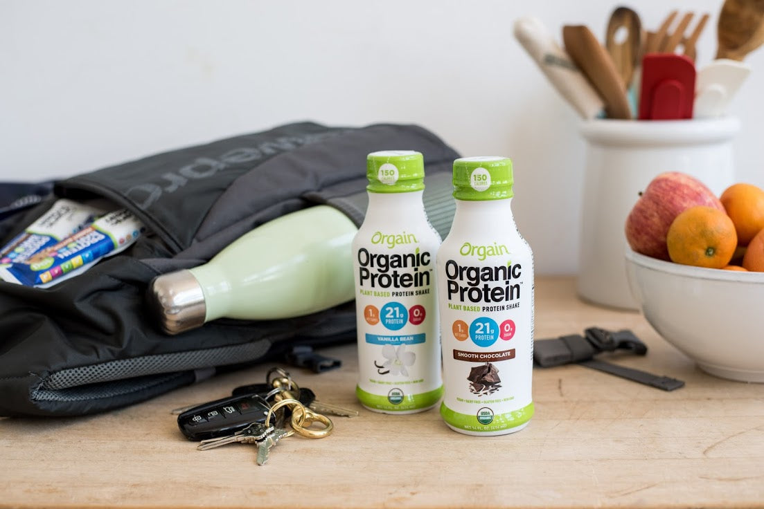 A wooden table holds a variety of items including a backpack, a water bottle, keys, an open protein bar, a bowl of mixed fruit, and two bottles of Orgain Organic Protein shakes in vanilla bean and creamy chocolate flavor.