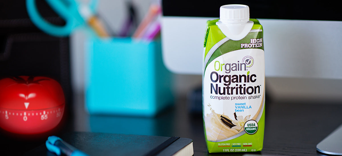 A carton of Orgain Organic Nutrition protein shake in Sweet Vanilla Bean flavor is placed on a desk. The carton is white and green with the product name and label visible. In the background, there are office supplies, a red timer, and a computer monitor.