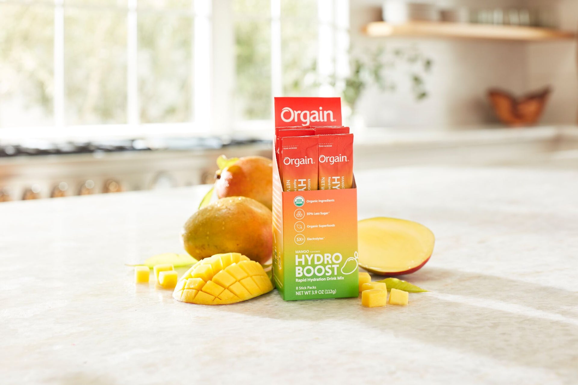 A picture of Orgain Hydro Boost electrolyte drink mix packets leaning against a sliced mango on a kitchen countertop. The packets are bright orange with white text, and the background features a bright, sunlit kitchen with a blurred window view.