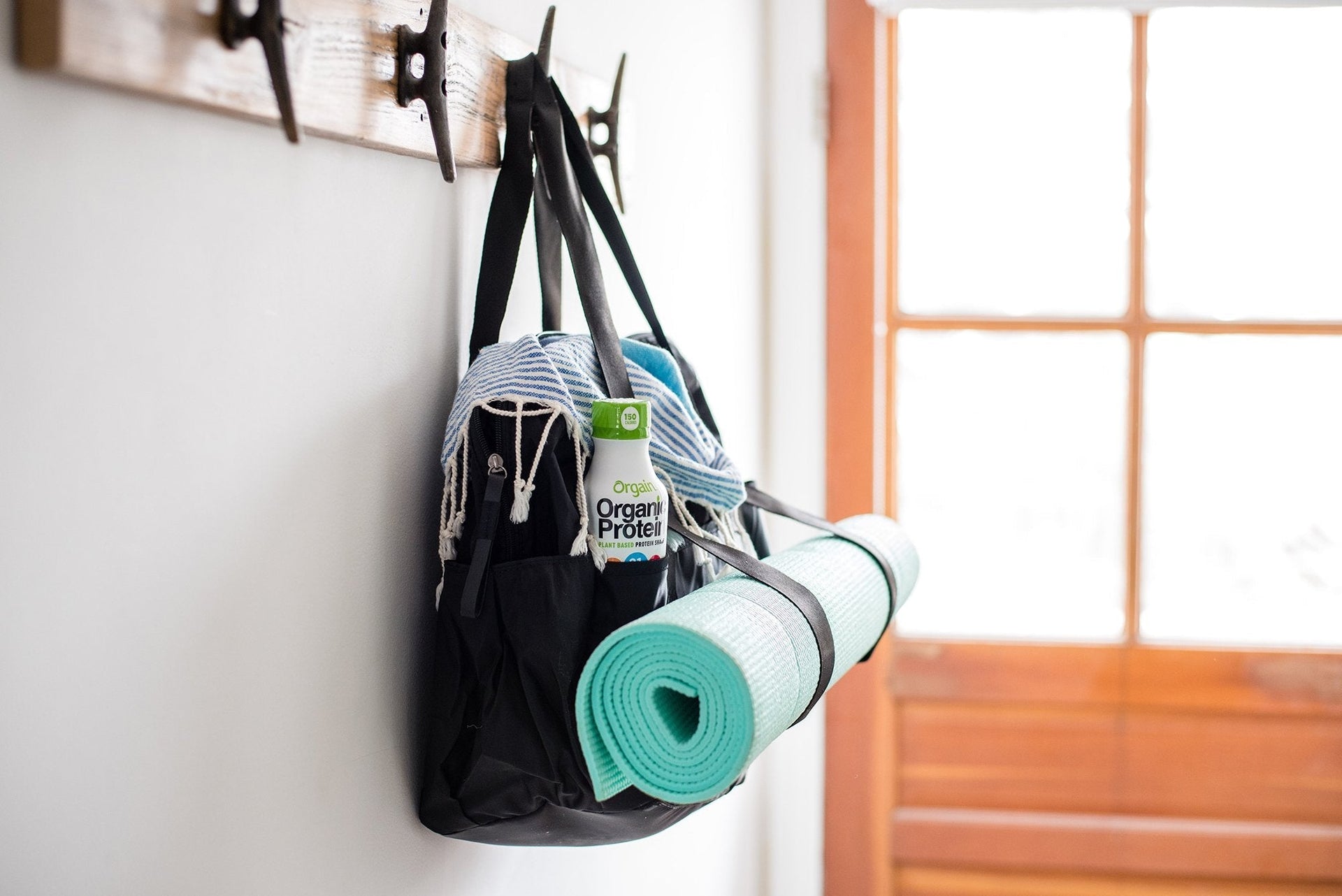 A black bag hangs on a wooden wall hook next to a glass door. The bag holds a rolled-up teal yoga mat, a blue-and-white striped towel, and a container of organic protein powder. The scene suggests a ready-to-go kit for a yoga session or gym workout.