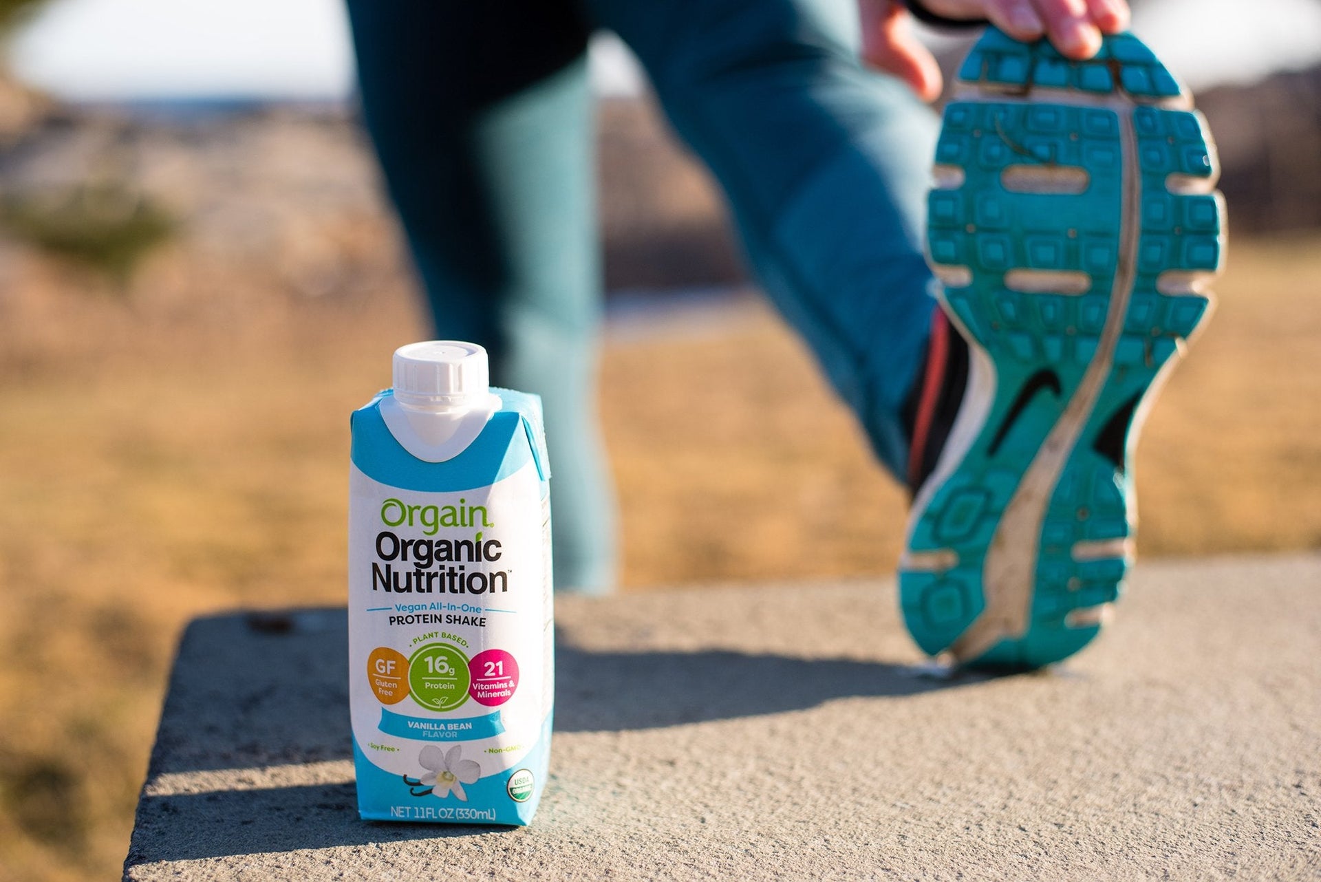 A person stretches their leg on a concrete surface outdoors. In the foreground, there is a container of Orgain Organic Nutrition protein shake in Vanilla Bean flavor. The person is wearing blue athletic clothing and turquoise running shoes.