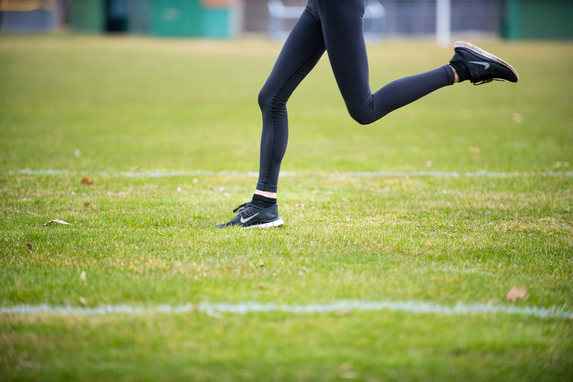 A person in black leggings and black running shoes is mid-stride, running on a grassy field. The background is blurred, focusing on the runner's legs and the green grass. White lines are visible on the grass, indicating a sports field.