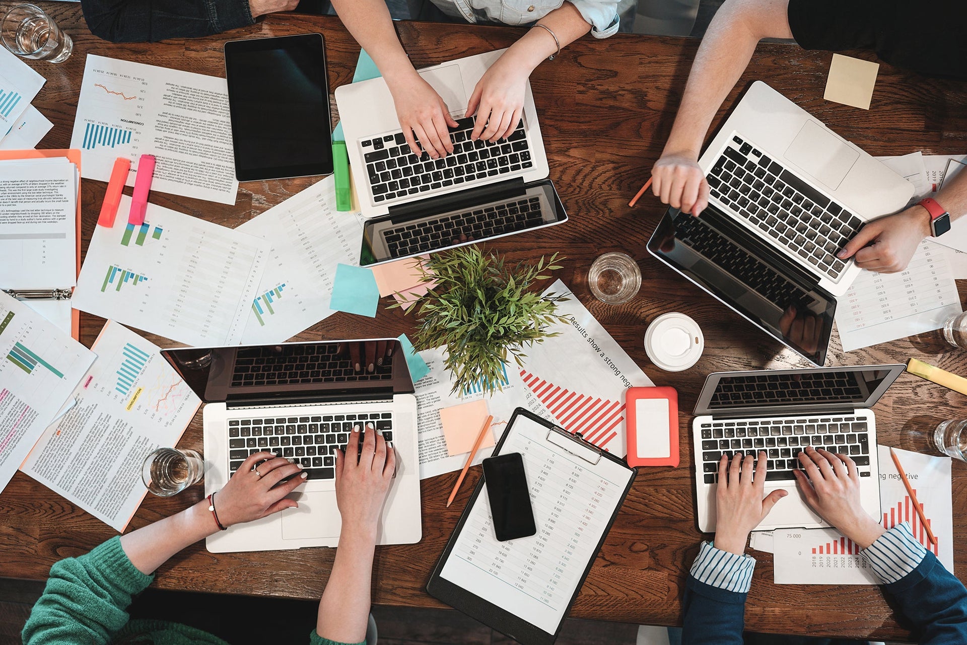 A group of people working together at a wooden table with laptops, documents, graphs, sticky notes, smartphones, and a small potted plant. They are focused and actively collaborating, with their hands visible on keyboards and papers.