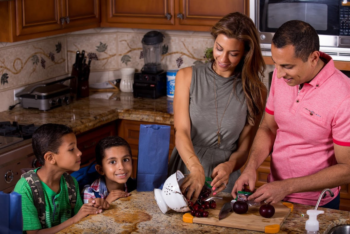 A family gathers around a kitchen island. A woman and a man prepare fruits together while two children, a boy in a green shirt and another in a blue shirt, watch attentively. The kitchen features wooden cabinets, a blender, and a tile backsplash.