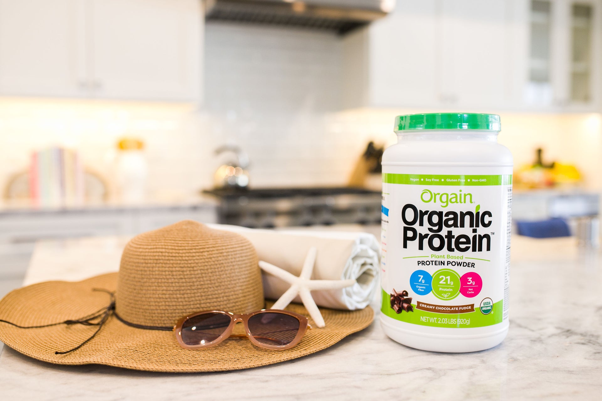 A jar of Orgain Organic Protein Powder is displayed on a kitchen counter. Beside it are a brown sun hat, a pair of sunglasses, and a white starfish with a rolled-up towel, indicating a summer or beach theme. The kitchen background is slightly blurred.