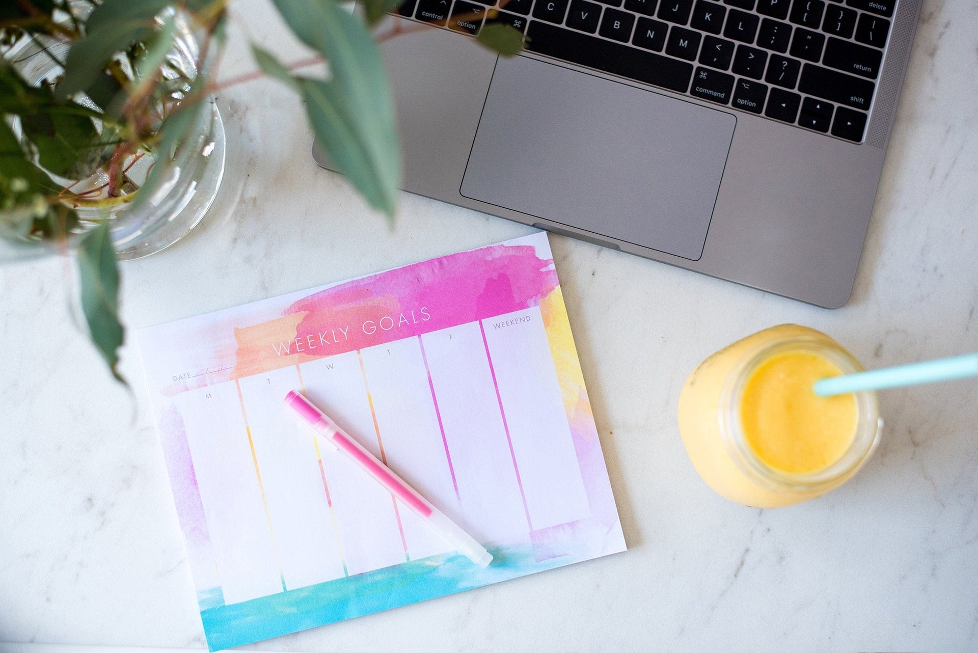 Overhead view of a workspace featuring a laptop, a weekly goals planner with a pen, a plant, and a mason jar with a yellow drink and a straw, all on a marble surface. The planner has vibrant colors at the top and is mostly empty.