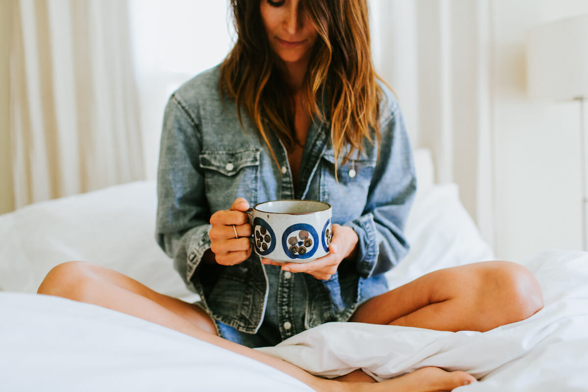 A woman sits cross-legged on a bed with white sheets, holding a patterned mug. She is dressed in a denim shirt and has long, wavy hair partially covering her face. The background is softly lit, creating a cozy and relaxed atmosphere.