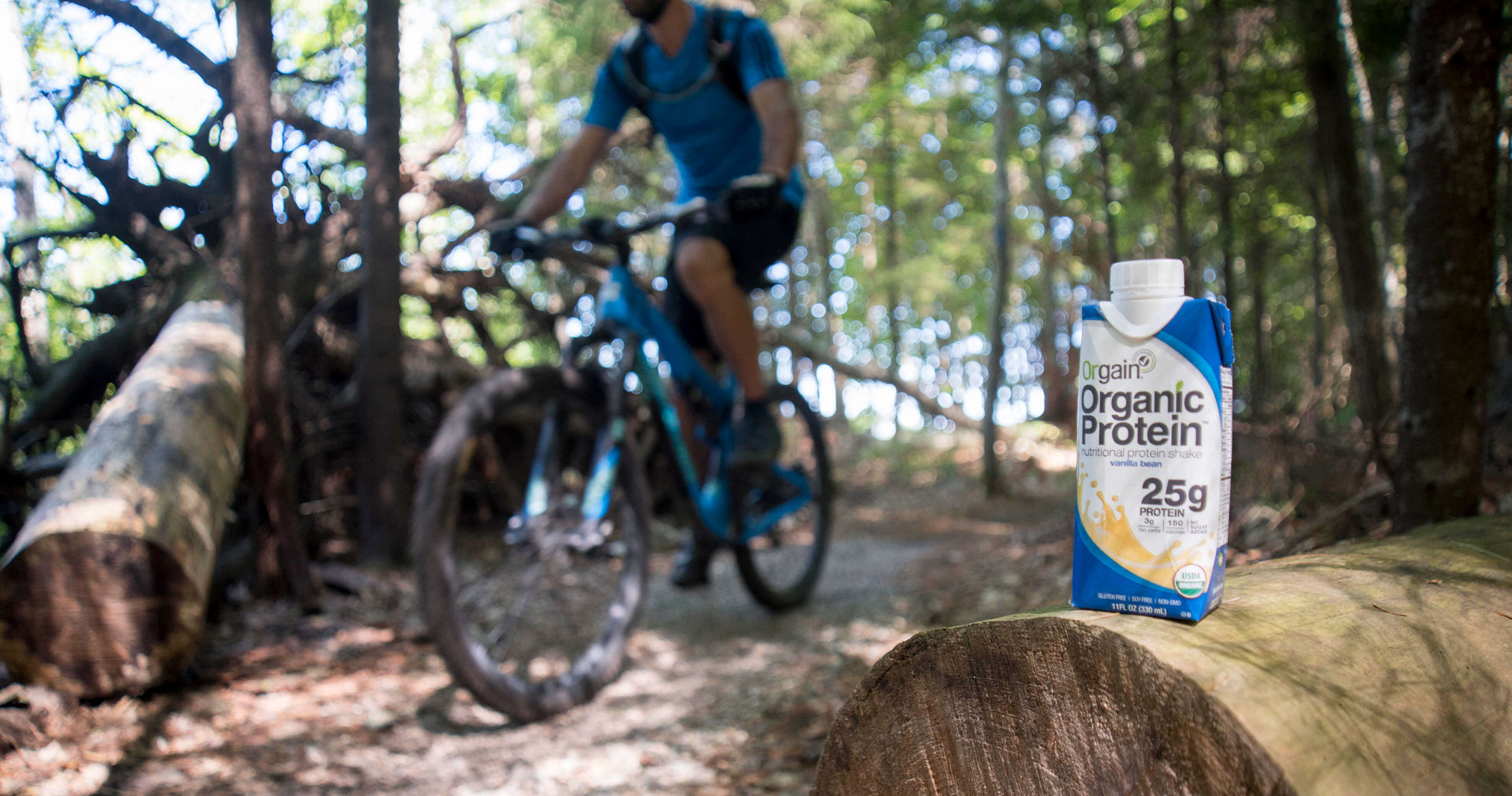 A cyclist rides through a forest trail. A container of Orgain Organic Protein shake is placed on a log in the foreground.