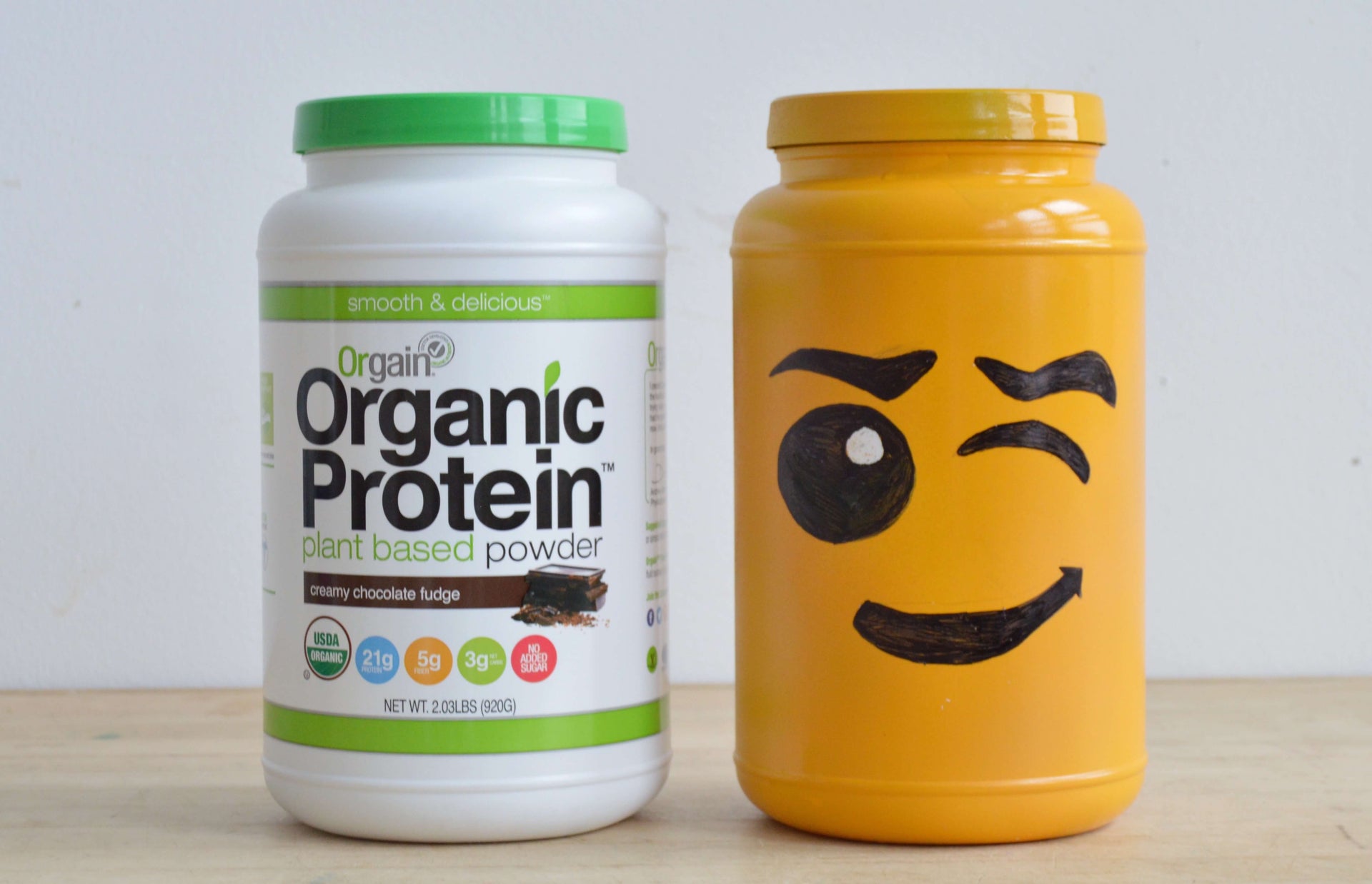 Two containers: a white jar labeled "Orgain Organic Protein Plant Based Powder" (creamy chocolate fudge flavor) and a yellow jar with a winking face, on a wooden surface.