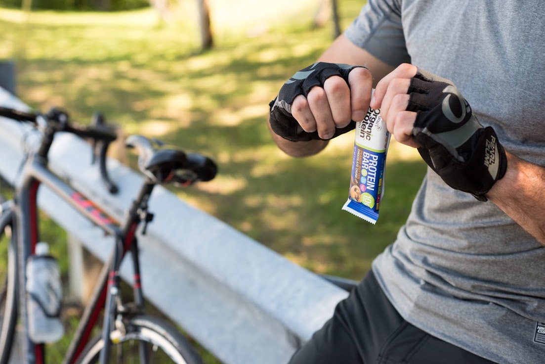 A cyclist wearing gloves and a gray shirt is opening a protein bar next to a parked bicycle on a sunny day. The background features green grass, trees, and a metal guardrail, giving the scene a relaxed, outdoor feel.