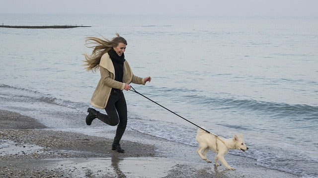 A woman with long hair, wearing a beige coat and black pants, is running and smiling while holding the leash of a small white dog on a beach. The sea is calm in the background, and it is a cloudy day.