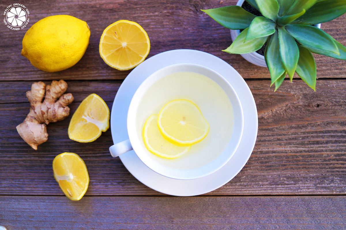 A white cup filled with a lemon-infused beverage on a saucer, containing lemon slices. Surrounding the cup are fresh lemon halves, ginger root, and a small potted succulent, all placed on a wooden surface.