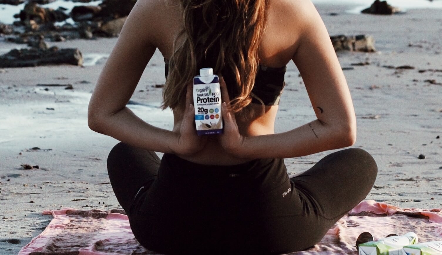 A person in a black sports bra and leggings sits cross-legged on a pink yoga mat at the beach, holding a shake bottle.