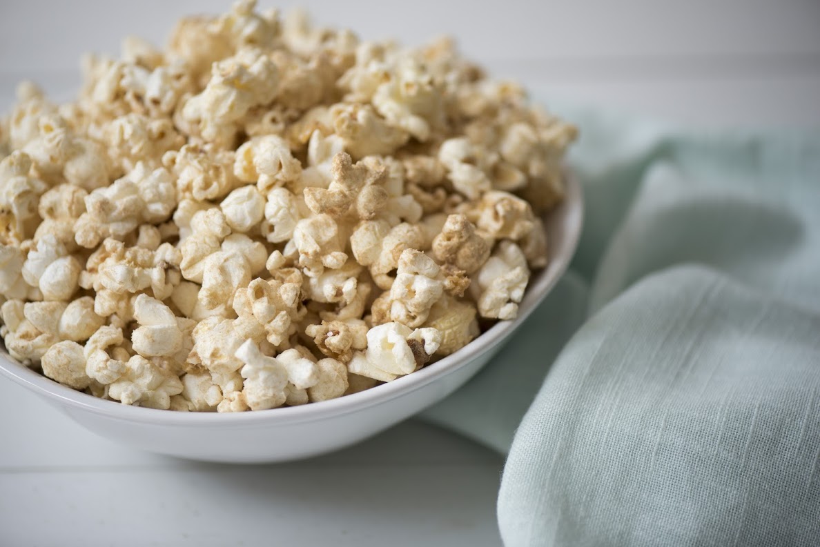 A white bowl filled with freshly popped popcorn is placed on a white surface. Next to the bowl, there is a light blue cloth napkin, slightly wrinkled, adding a touch of color to the scene. The background is simple and soft-focused.