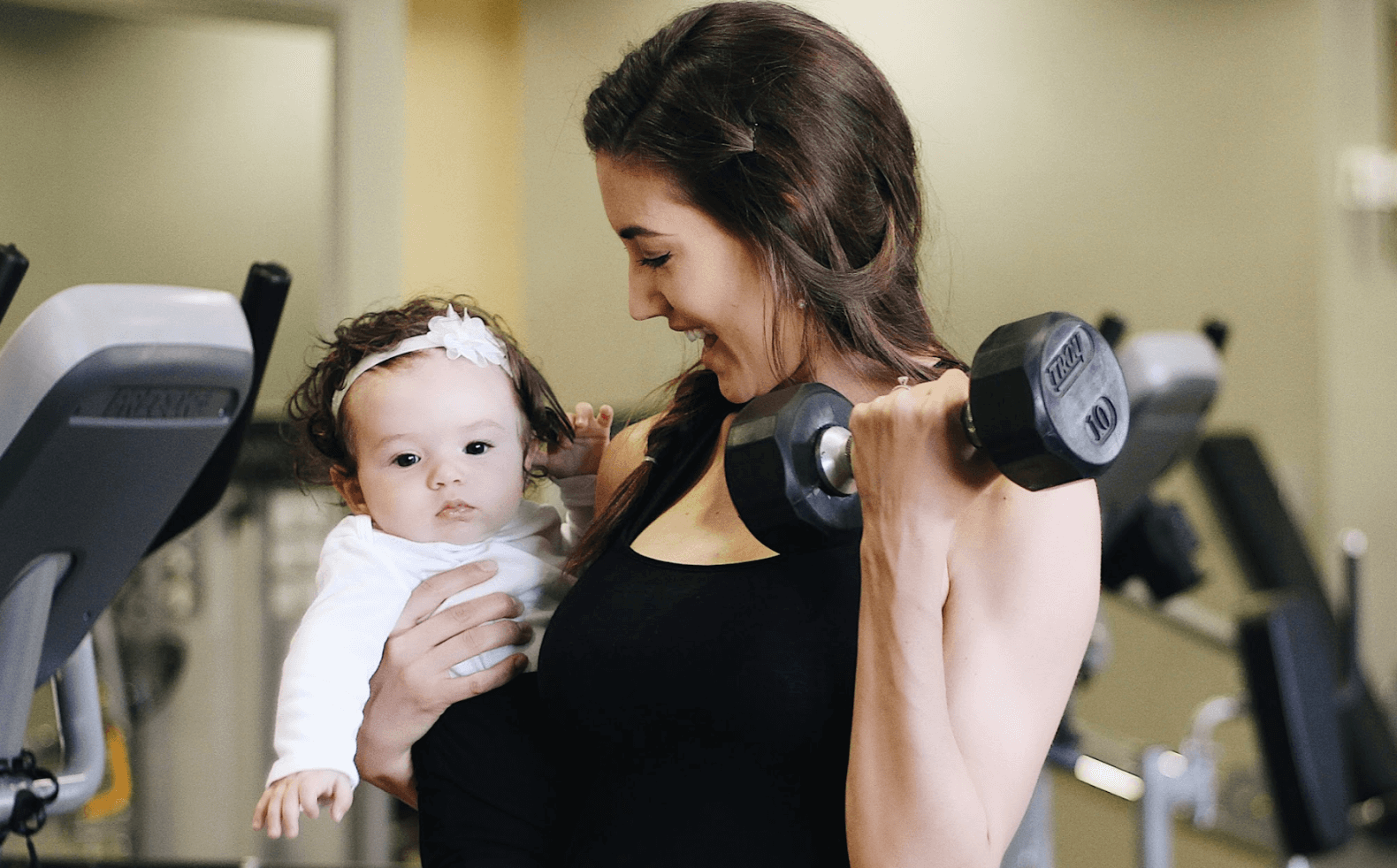 A woman holding a baby in her left arm lifts a dumbbell with her right hand while smiling. She is in a gym, surrounded by fitness equipment. The baby, wearing a white outfit and a headband with a bow, looks into the distance.