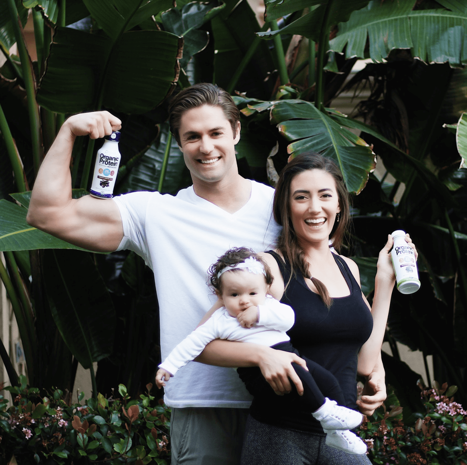 A smiling man flexes his arm and holds a protein shake bottle, while a woman beside him holds a baby and a shake bottle.