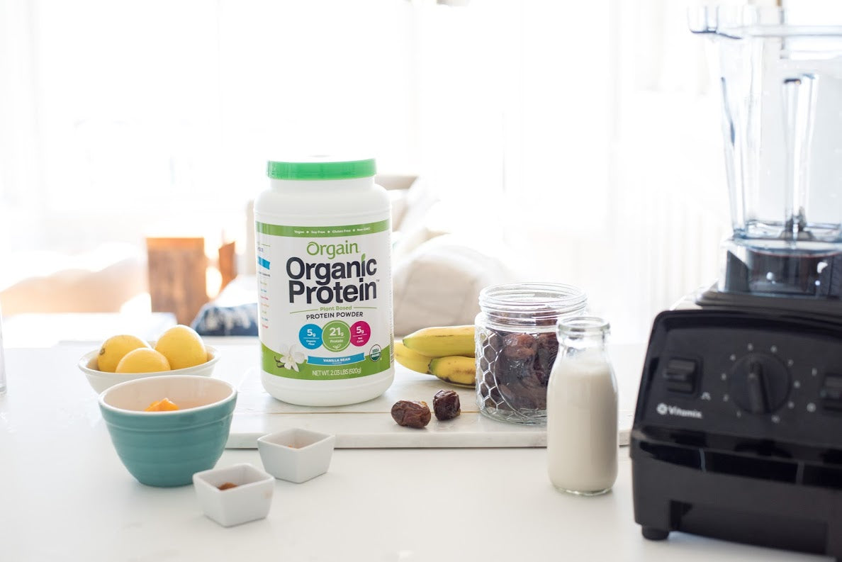 A tub or Orgain's Organic Protein and a bunch of other smoothie ingredients beside a blender.