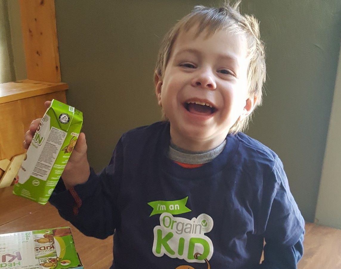 A child with a big smile is holding a small box in one hand and wearing a shirt that reads "I'm an Orgain Kid." The child has short hair and is sitting indoors, with a greenish-brown wall in the background.