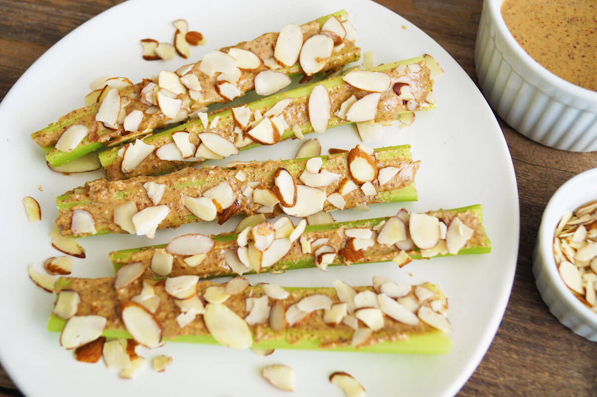 A white plate holds several celery sticks topped with almond butter and sprinkled with sliced almonds. A small bowl of almond butter and a ramekin containing additional sliced almonds are placed beside the plate on a wooden surface.