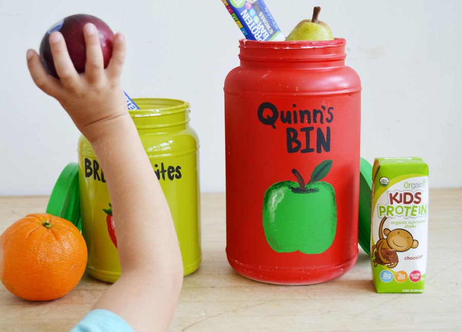 A child's hand reaches for an apple beside brightly painted containers labeled "Quinn's Bin" with an apple graphic, and "Brinley's Bites" with a carrot graphic. An orange, a pear, and an Organix Kids Protein chocolate drink are also on the table.