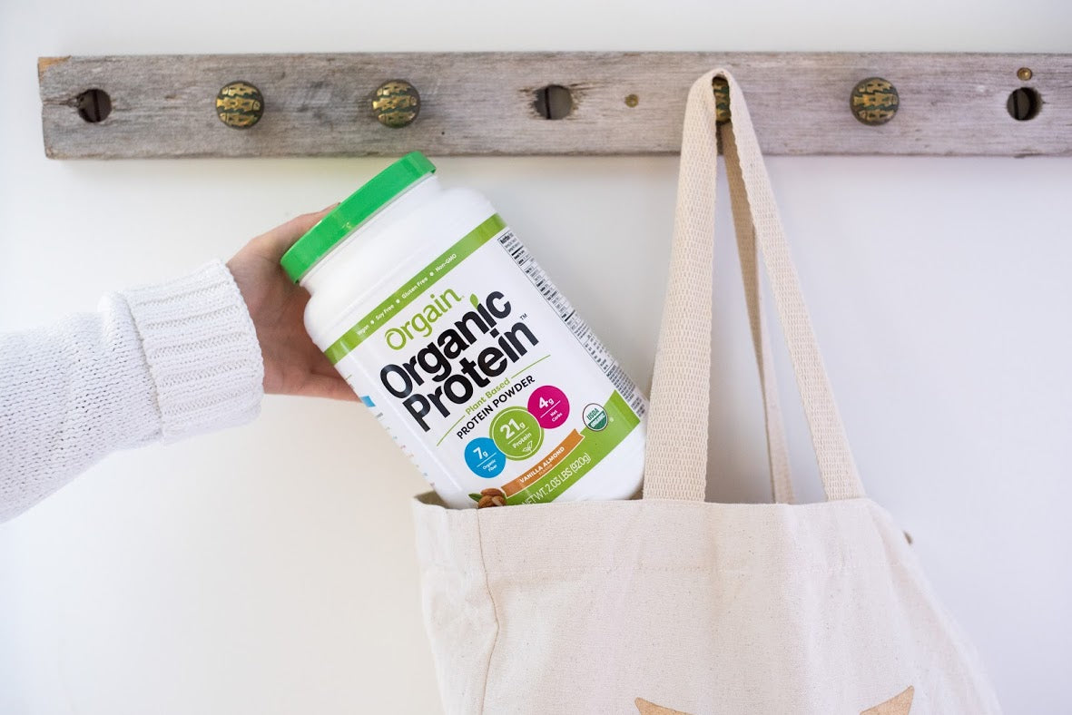 A woman sliding a tub of Orgain's Organic Protein powder into a canvas bag hanging on a coatrack