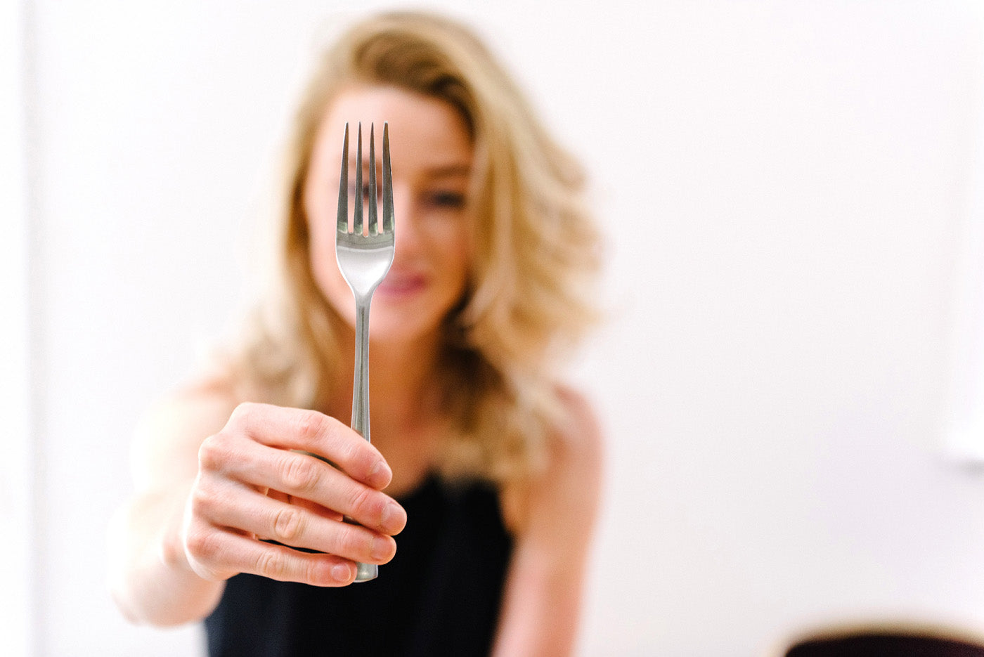 A person with long, wavy blonde hair is holding a fork toward the camera, slightly out of focus. The background is a plain, white wall, and the person is wearing a black top.