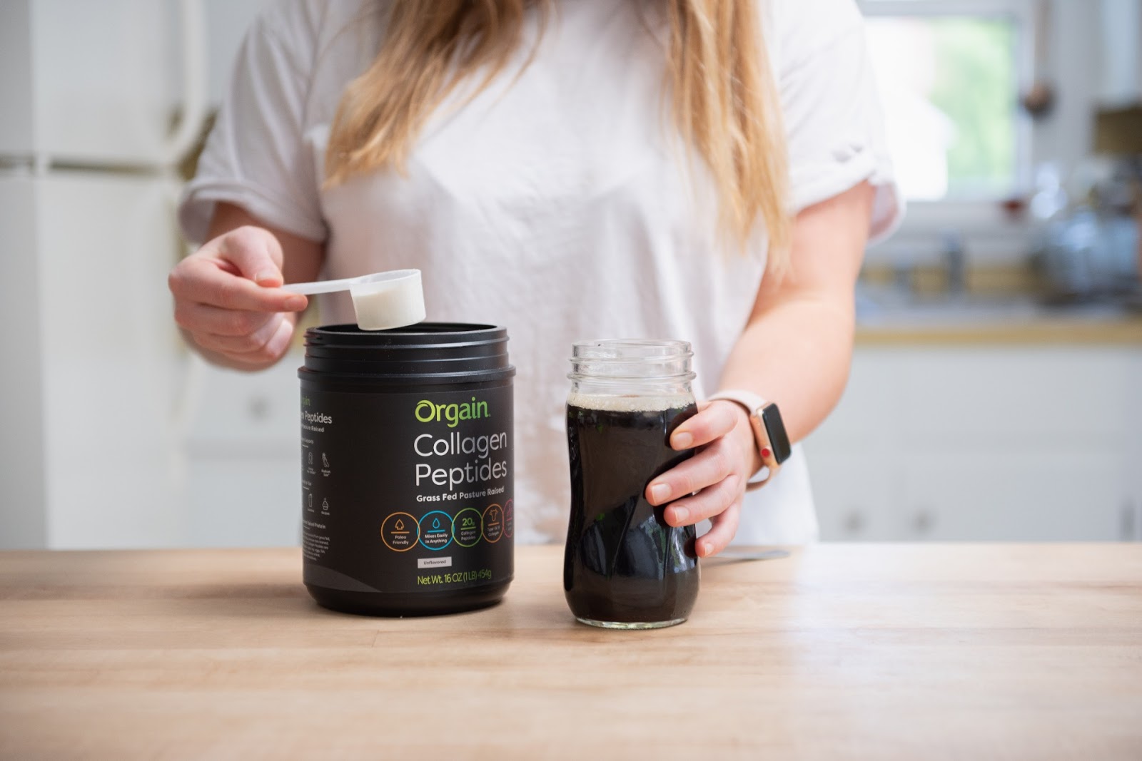 A person with long blond hair, wearing a white T-shirt, scoops collagen peptides powder from a black Orgain container with a white scoop. They are holding a glass jar filled with a dark liquid on a wooden table in a kitchen.