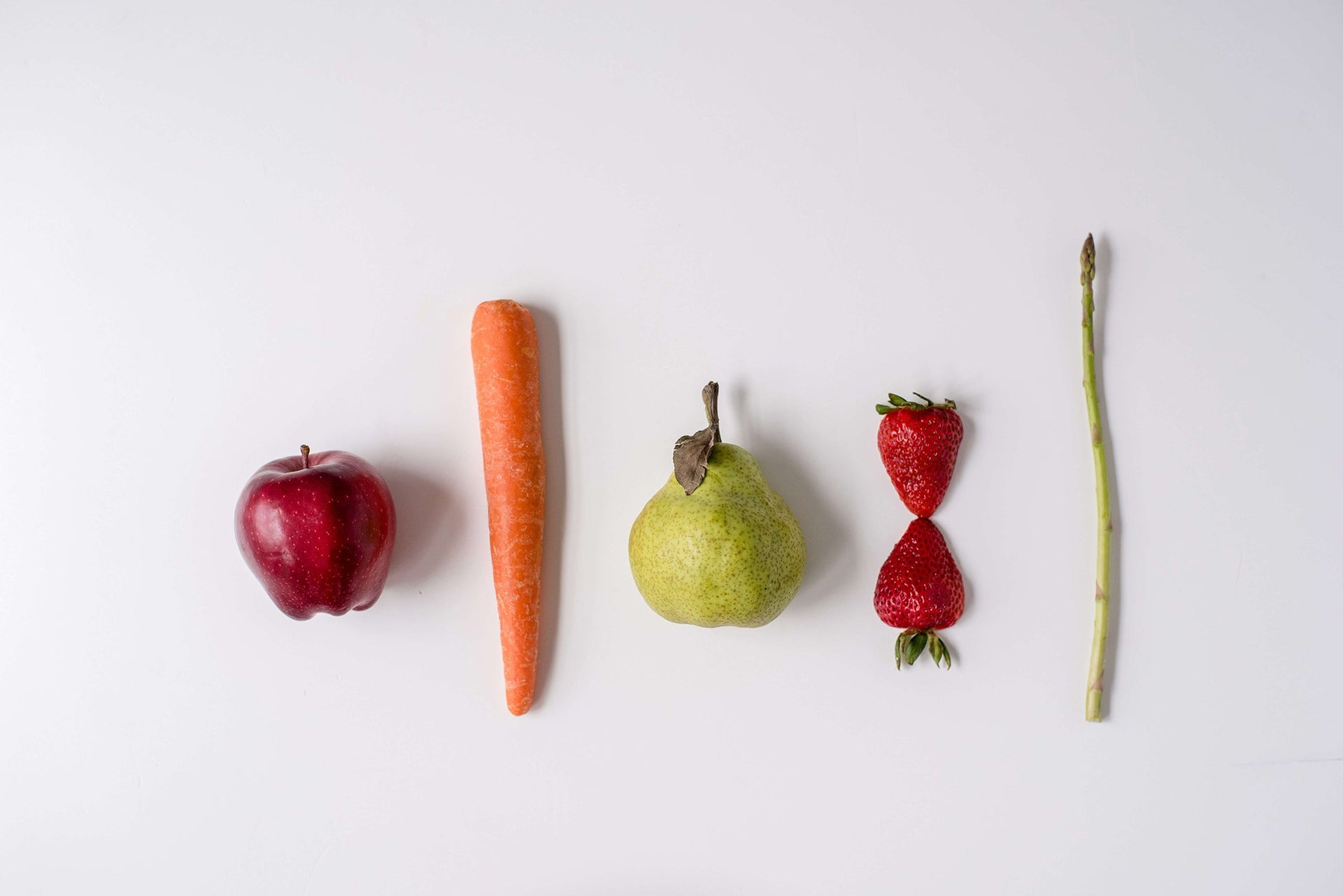 A lineup of fruits and vegetables placed in a row on a white background. From left to right: a red apple, an orange carrot, a green pear, two strawberries stacked one on top of the other, and a green asparagus spear.