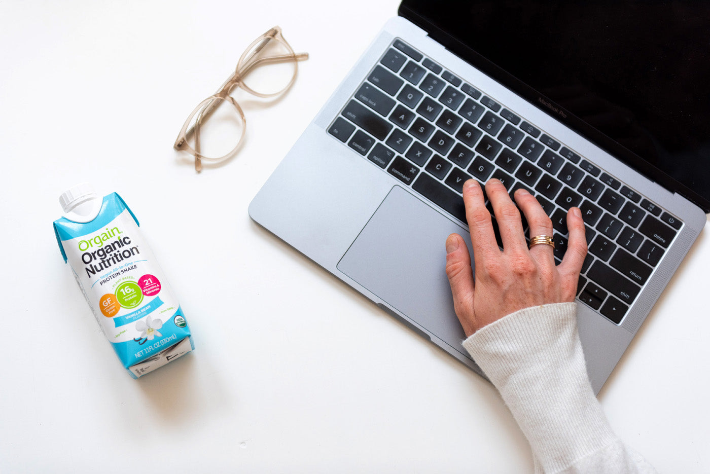 A person wearing a white long-sleeve shirt is using a laptop with their right hand on the keyboard. To the left of the laptop, there is a pair of eyeglasses and a carton of Orgain Organic Nutrition shake on a white surface.