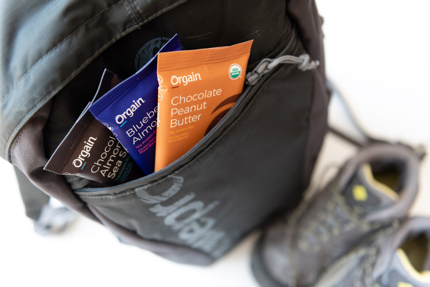 A black backpack with Orgain protein bar packages visible in the front pocket. The bars are in different flavors: Chocolate Almond Sea Salt, Blueberry Almond, and Chocolate Peanut Butter. A pair of hiking shoes is placed next to the backpack.