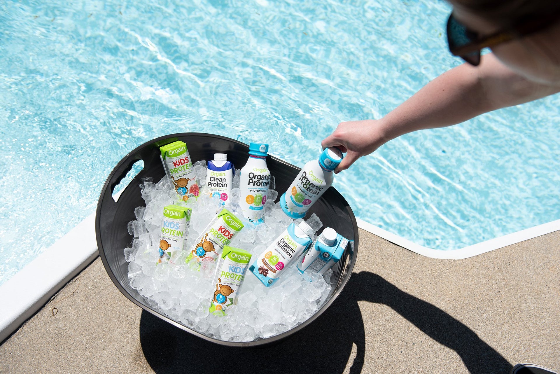 A person is reaching for a protein drink from a black bucket filled with ice and various protein drinks. The bucket is placed on the edge of a concrete surface next to a clear blue swimming pool.