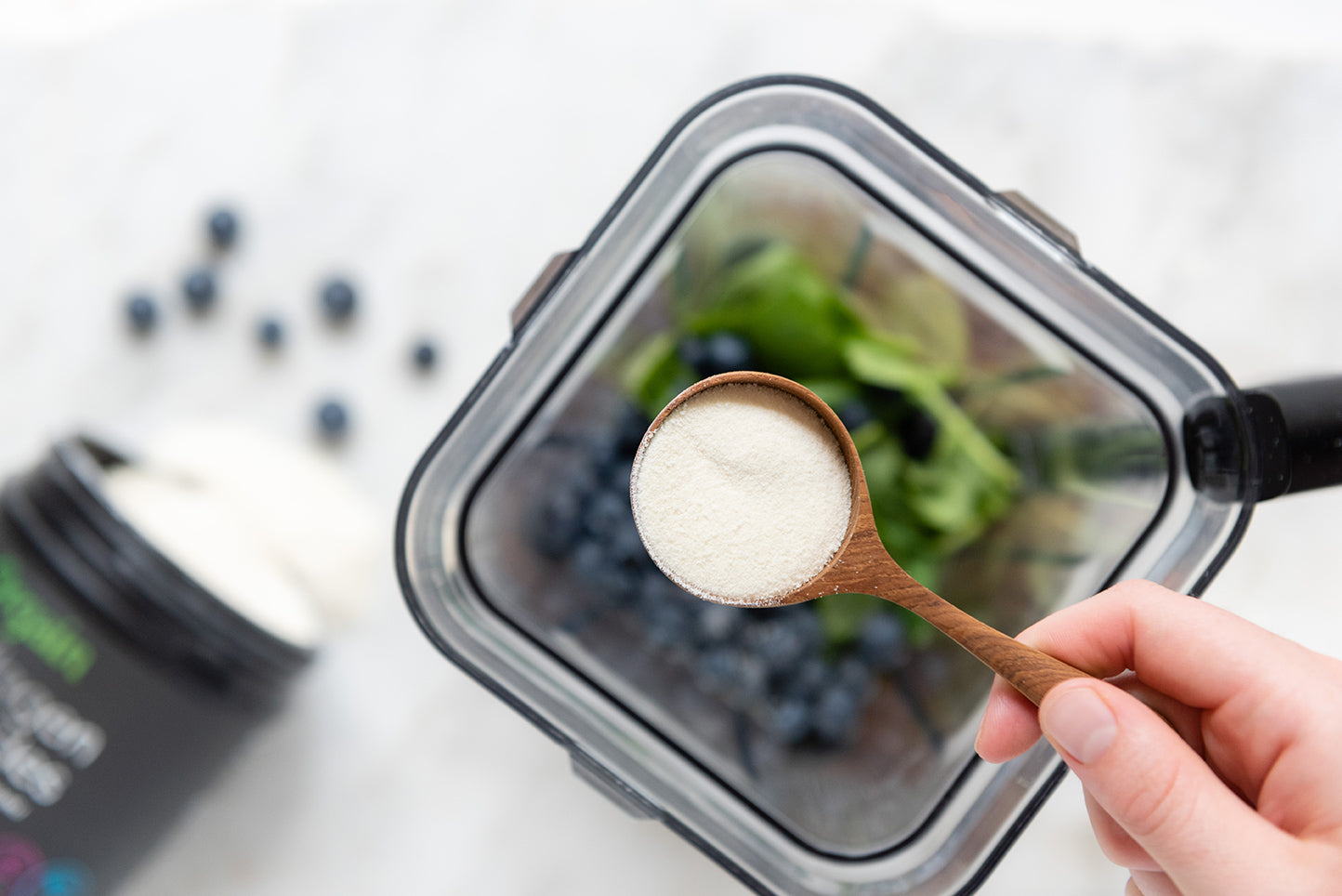 A hand holding a wooden spoon with a scoop of white powder above an open blender. Inside the blender are leafy greens and blueberries. A container of protein powder is on the countertop next to a few scattered blueberries.