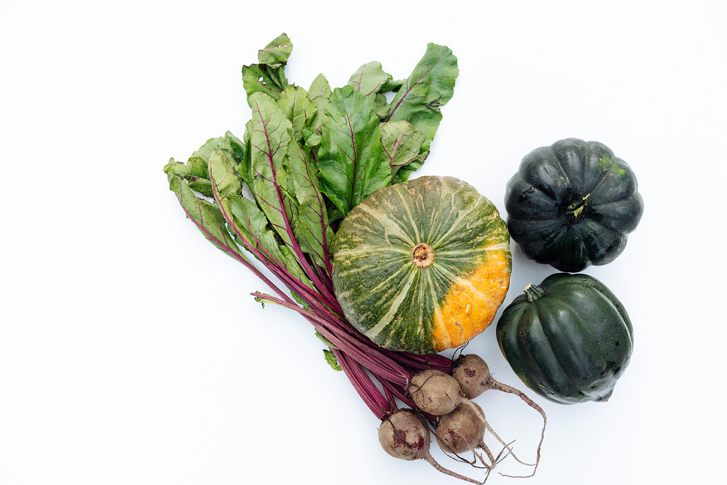 A bunch of fresh vegetables against a white background. The vegetables include a mix of leafy beetroot greens with attached beetroots, a green and orange striped pumpkin, and two dark green acorn squashes.