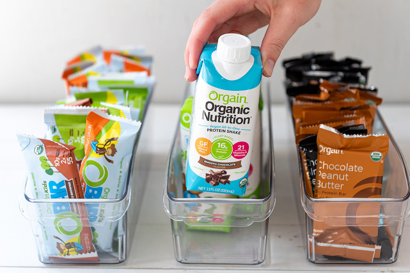 A hand holding an Orgain Organic Nutrition protein shake container above a clear bin filled with more containers. There are two adjacent bins, one containing Orgain protein bars in green wrappers and another with similar bars in brown wrappers.

.