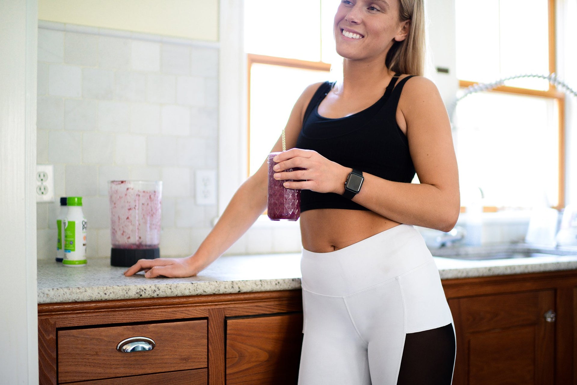 A woman in a black sports bra and white leggings stands in a bright kitchen, holding a purple smoothie. She is smiling and leaning against a counter with a blender and a green bottle visible in the background. The kitchen has wooden cabinets and a window.