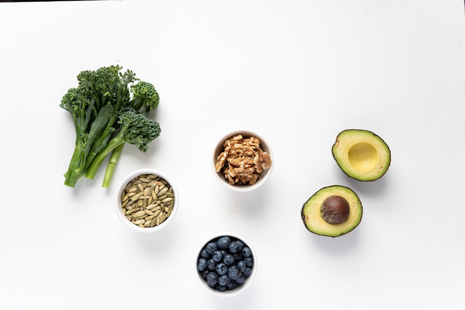 A variety of healthy foods are arranged on a white surface: broccoli florets, a cut avocado revealing its seed, a small bowl of walnuts, a small bowl of pumpkin seeds, and a small bowl of blueberries.