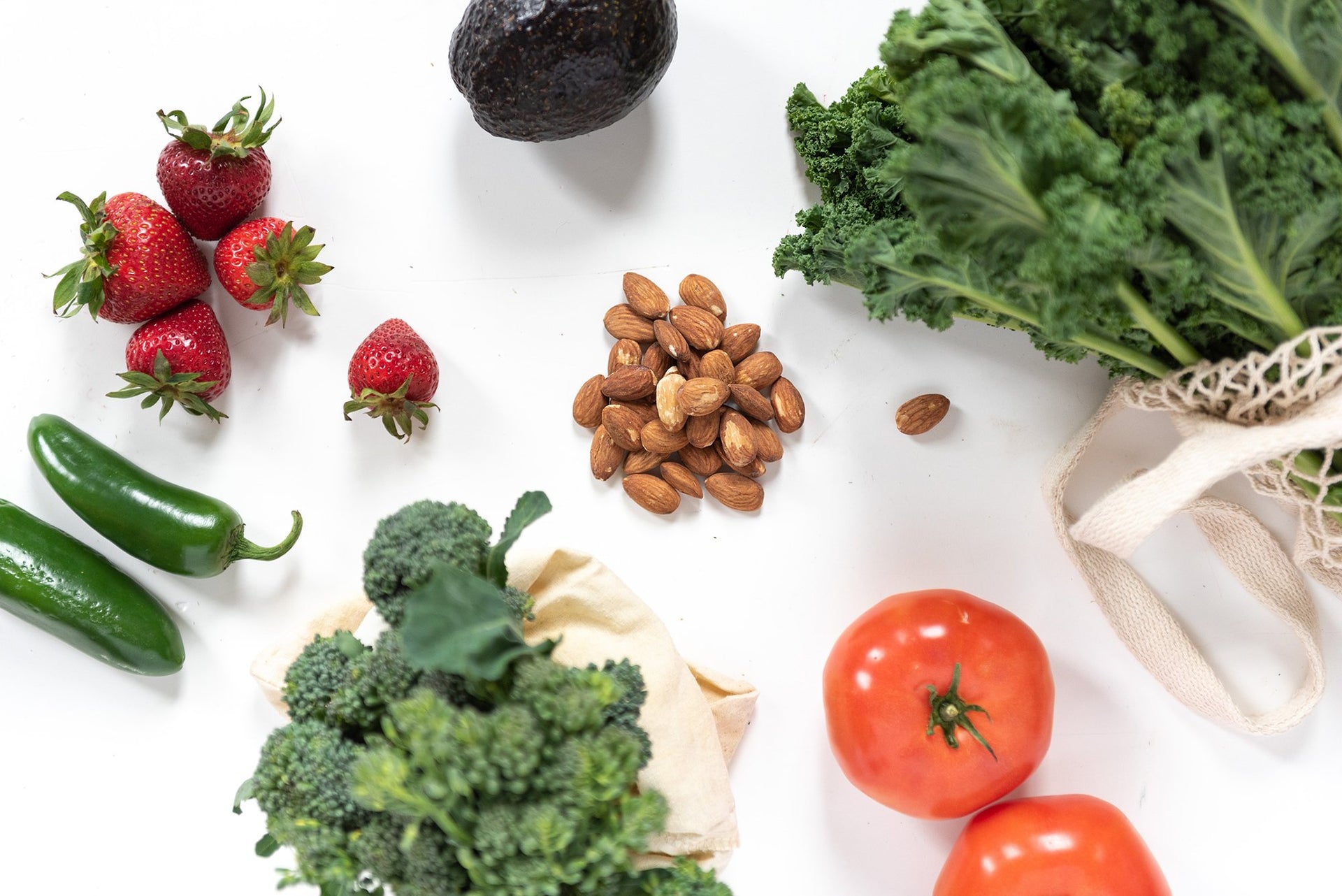 A collection of fresh produce and almonds on a white surface. Includes strawberries, avocado, jalape√±os, kale, broccoli, tomatoes, and a pile of almonds in the center. Some vegetables are placed in cloth bags and a reusable mesh bag.