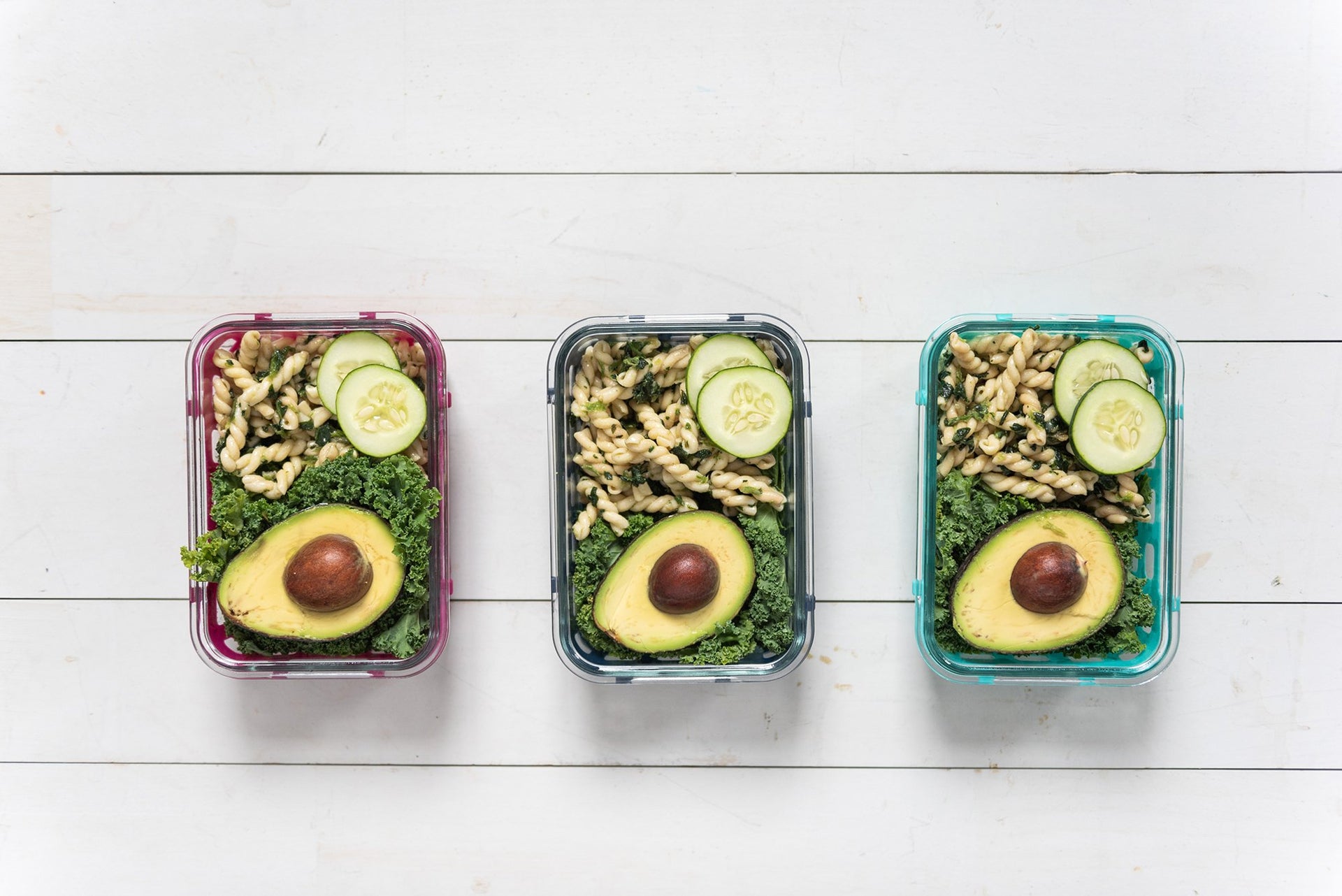 Three meal prep containers are lined up horizontally against a white wooden surface. Each container holds kale, half of an avocado, pasta salad, and two slices of cucumber, showcasing a colorful and healthy meal arrangement.