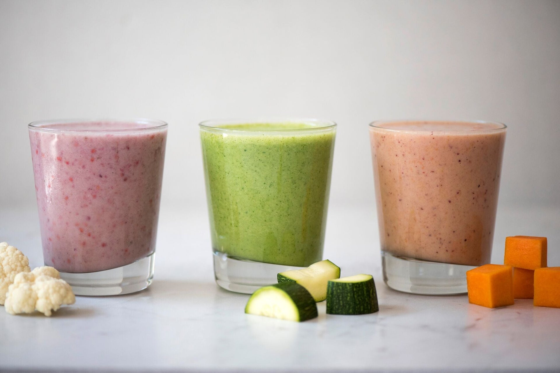 Three glasses of smoothies in different colors are lined up on a white surface. The left smoothie is pink, the middle one is green, and the right one is peach. Cauliflower, cucumber pieces, and diced squash are placed in front of the glasses.