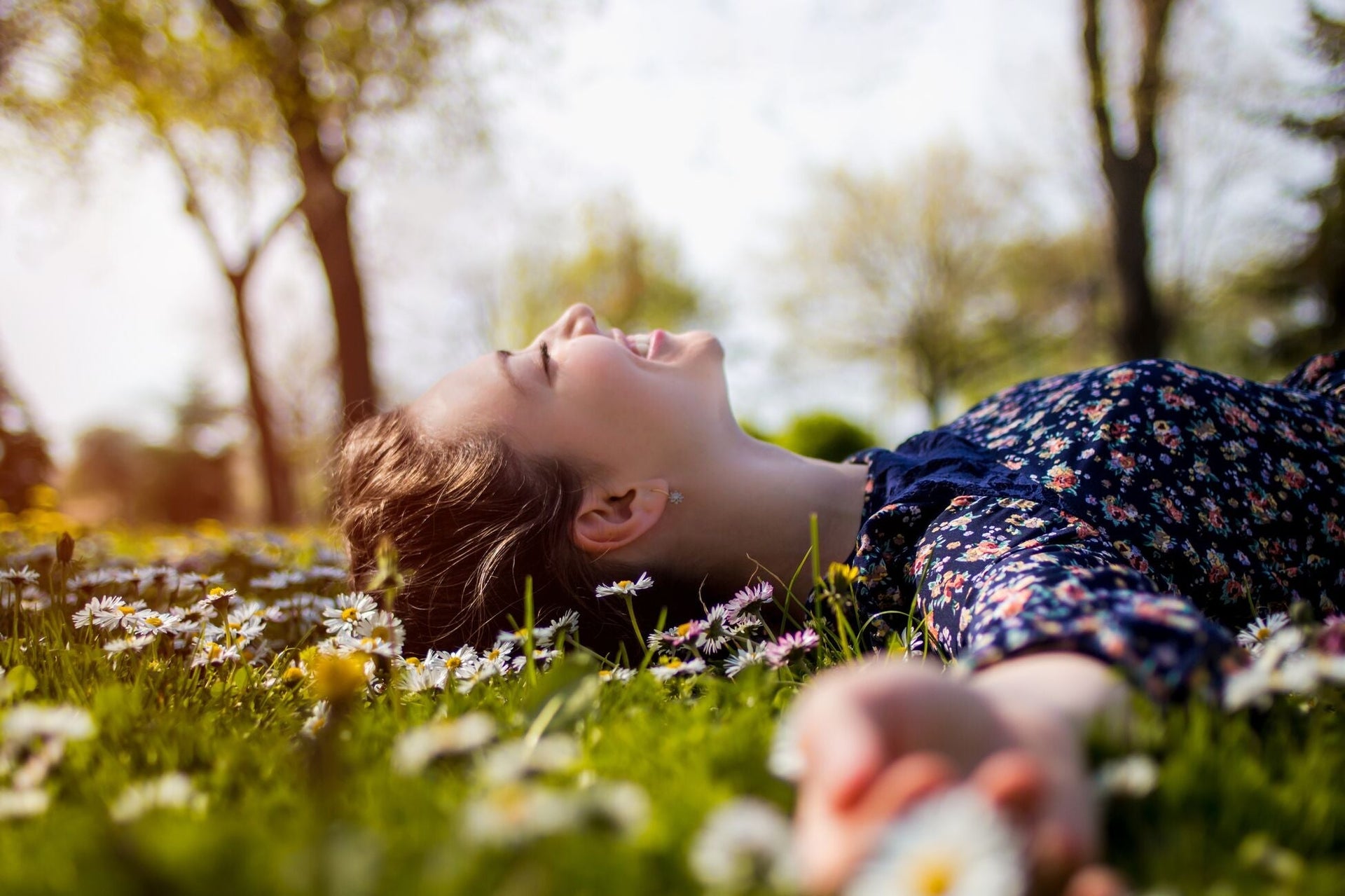 A young woman lies on grass with her eyes closed, smiling joyfully. She is surrounded by blooming daisies, wearing a floral dress. The background features trees and soft sunlight filtering through, creating a serene and happy atmosphere.