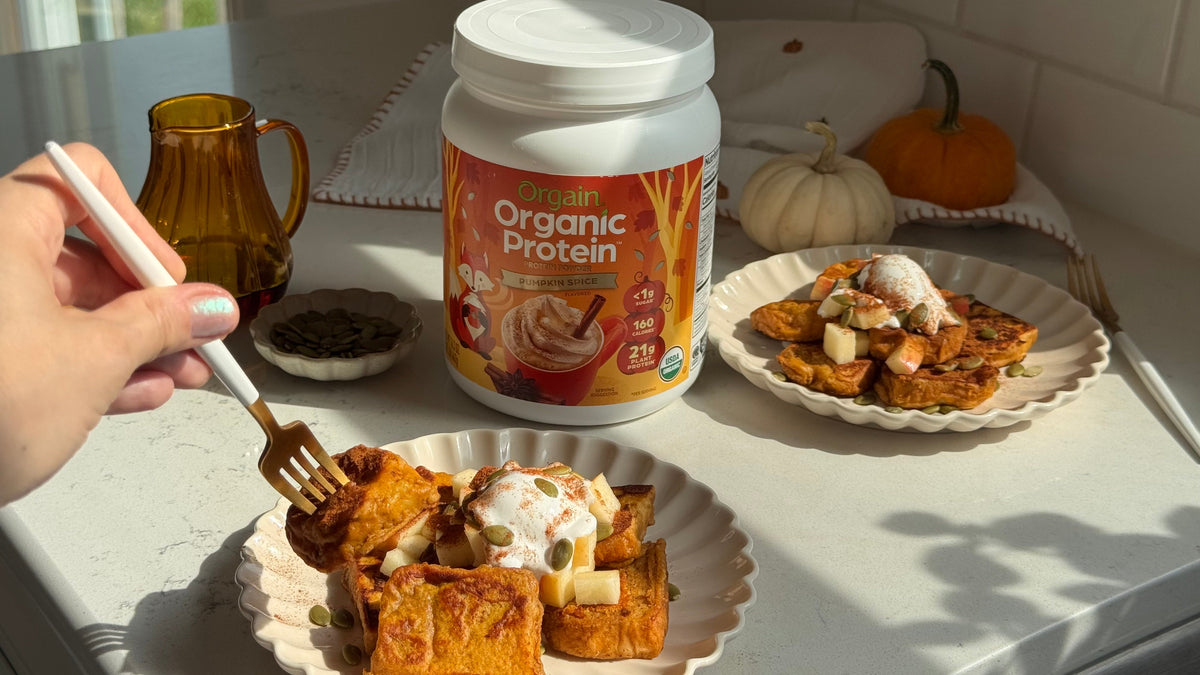 Orgain Organic Protein Powder in Pumpkin Spice beside two plates of pumpkin French toast topped with whipped cream, diced apple, pumpkin seeds, and cinnamon, with a hand holding a gold fork over one plate.