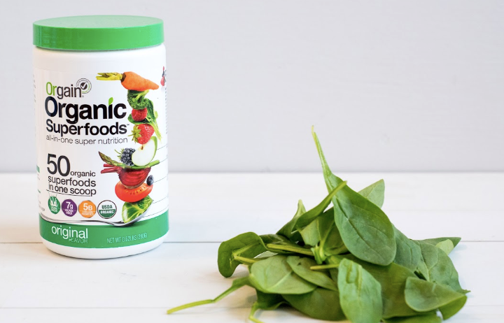 A white container of Orgain Organic Superfoods powder with a green lid is placed next to a small pile of fresh spinach leaves. The container's label states it contains 50 organic superfoods in one scoop and highlights its nutritional benefits.