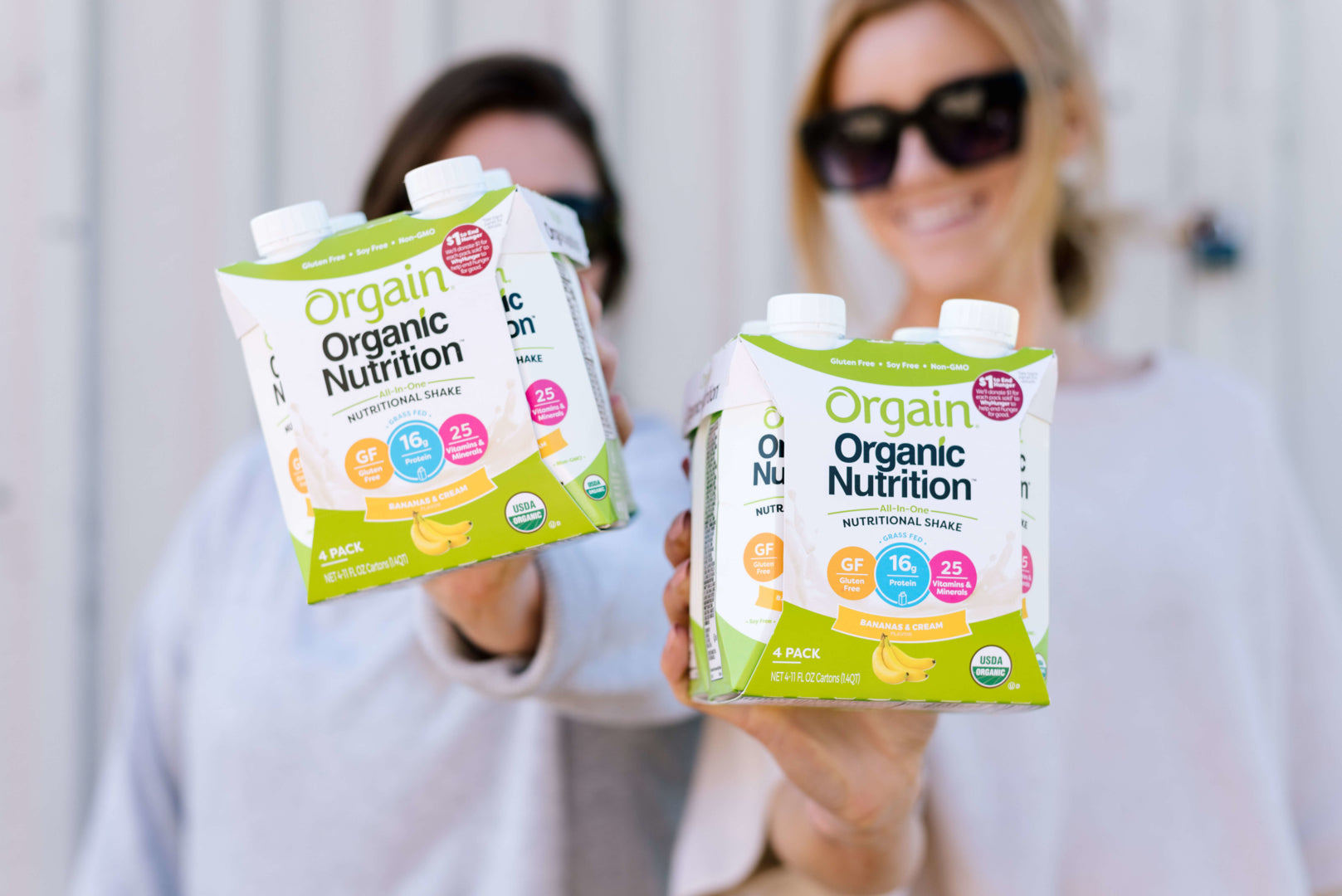 Two people holding packs of Orgain Organic Nutrition shakes towards the camera, highlighting banana flavor.