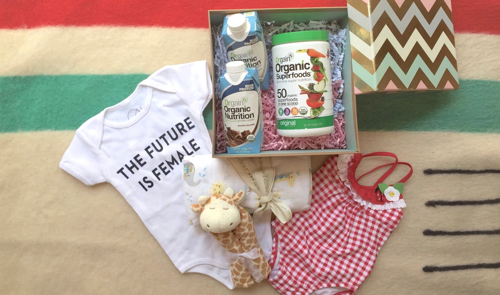 A gift box lies open on a colorful blanket, revealing items for a baby: a white onesie with the text "THE FUTURE IS FEMALE," a red and white checkered swimsuit, a plush giraffe toy, and organic nutrition products.