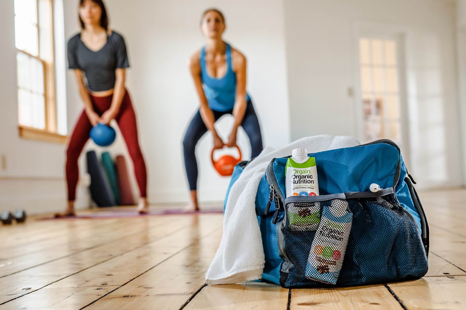 Two people engage in kettlebell exercises in a brightly lit room with wooden floors. In the foreground, a blue gym bag with a towel draped over it contains organic nutrition drinks and an apple. Yoga mats and gym equipment are visible in the background.