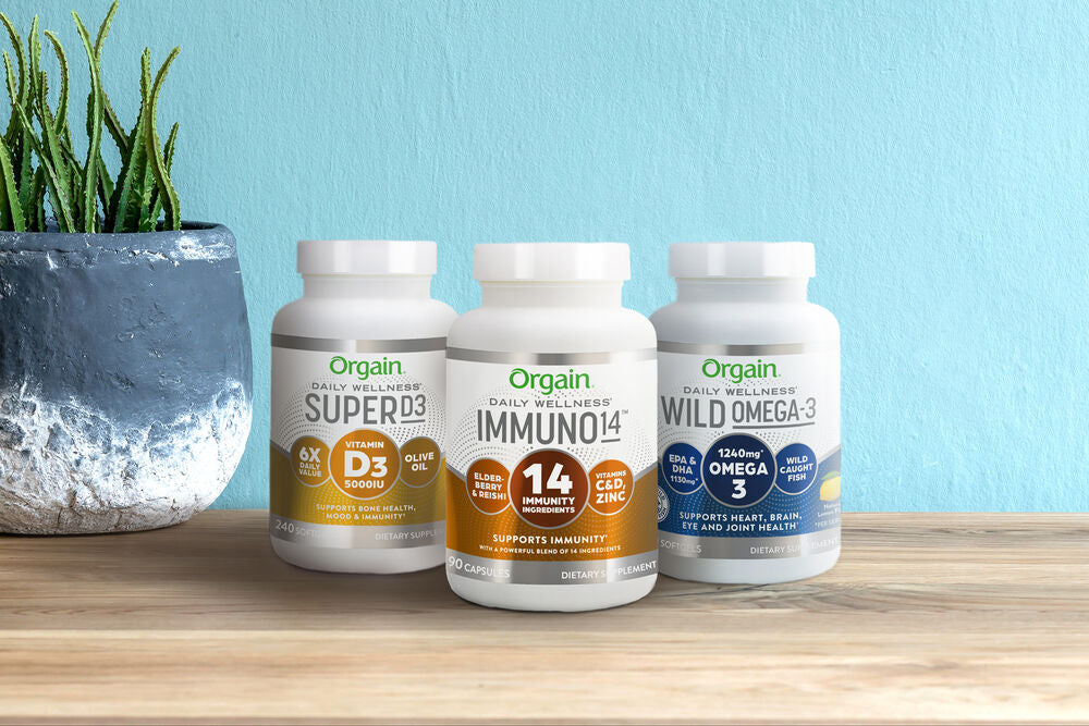 Three white containers of Orgain's health supplements are displayed on a wooden surface against a light blue background. From left to right, the products are labeled SUPERD3, IMMUNO14, and WILD OMEGA-3. A small potted plant is on the far left.