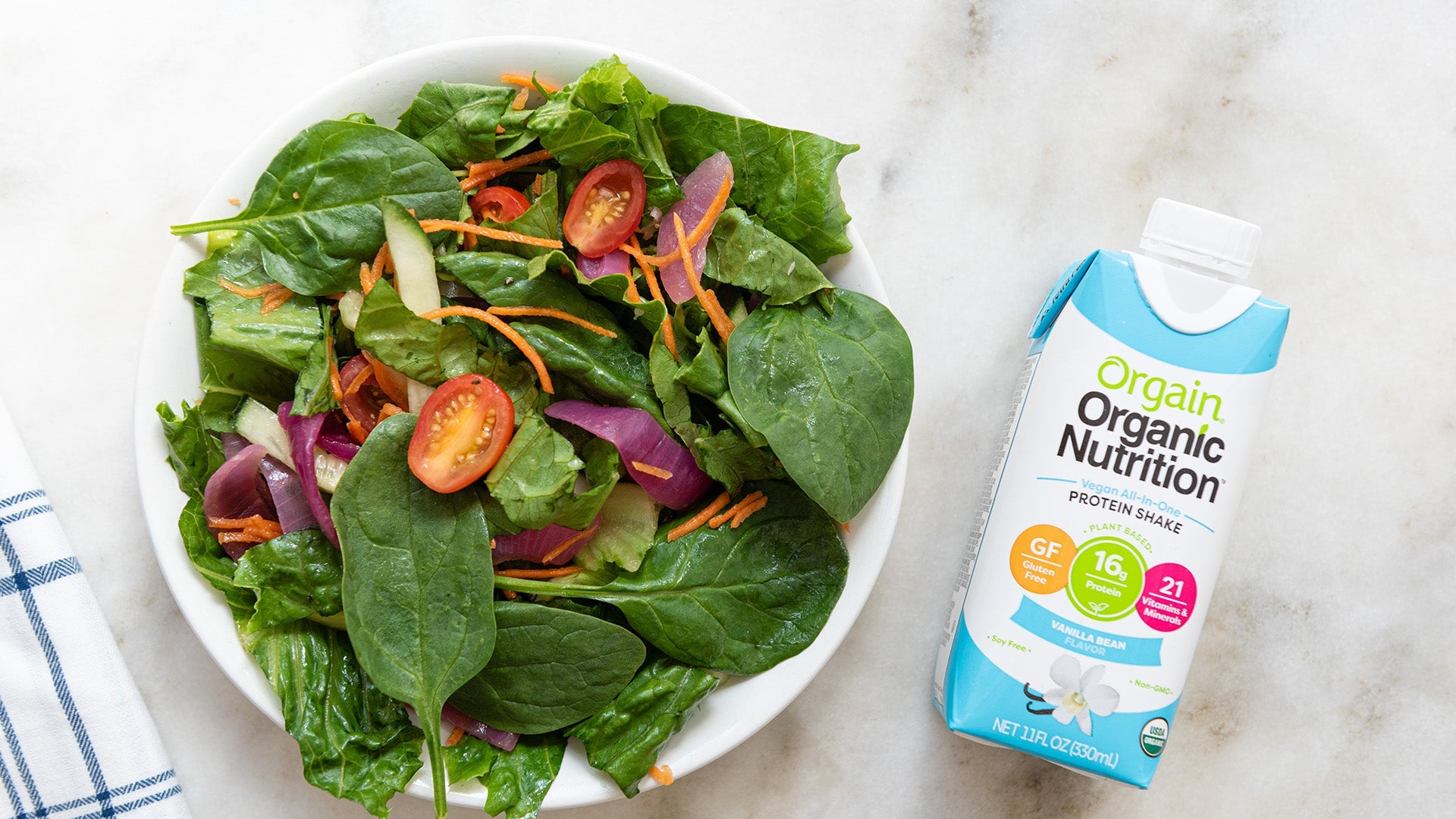 A fresh salad with spinach, carrots, cherry tomatoes, and onion in a bowl next to an Orgain Organic Nutrition shake carton.