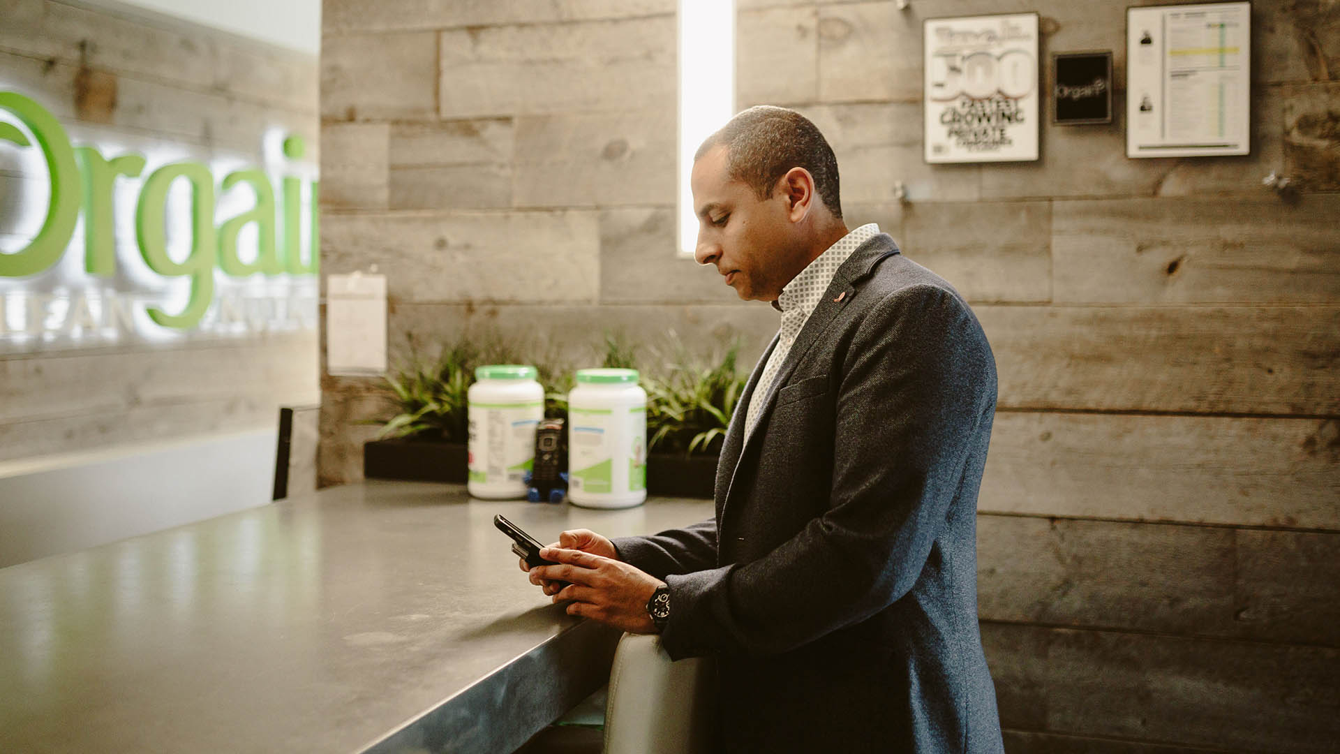 A man in a gray blazer stands at a counter, looking down at his phone. Behind him is a wall with wooden panels, green plants, and containers of protein powder. A partially visible sign reads "Orgain." Framed posters and certificates hang on the wall.