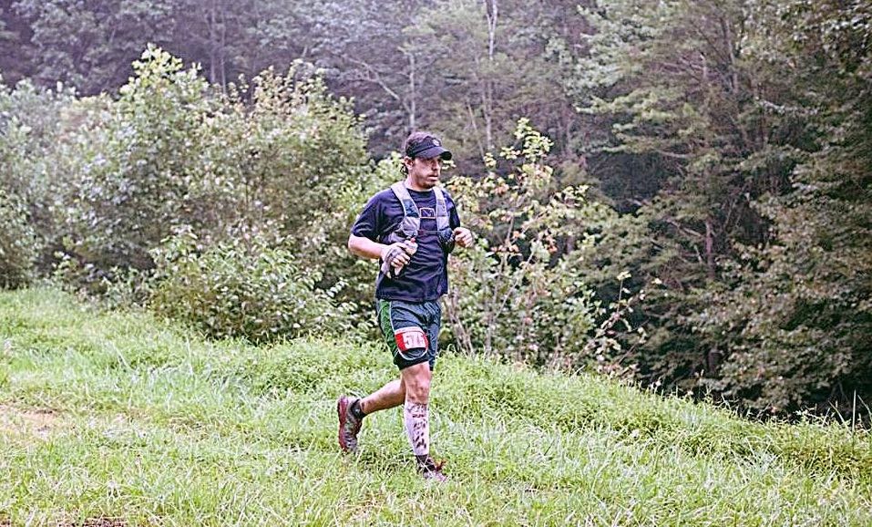 A person is running on a grassy trail surrounded by lush greenery and trees. They are wearing a hat, a vest, a number bib, and holding hiking poles. The environment looks calm and serene with fog in the background.