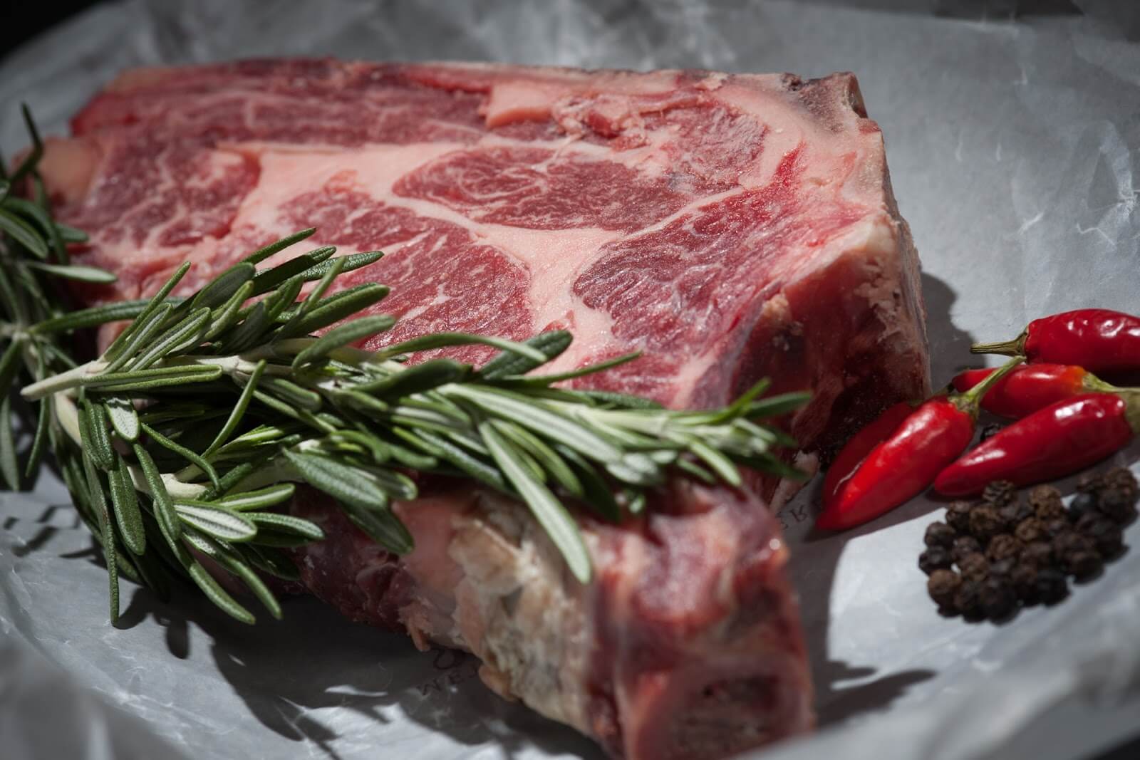 A raw marbled steak lies on white parchment paper. Fresh sprigs of rosemary and several red chili peppers are placed on top of the meat. A small cluster of black peppercorns is also on the paper next to the steak.